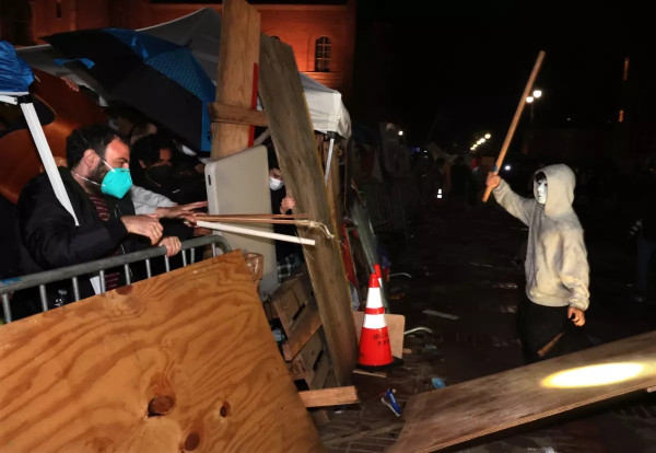 An outdoor scene at night showing a clash between masked individuals, some holding wooden planks and sticks. The background includes makeshift barricades made of wood and metal, with additional fencing and traffic cones. The atmosphere is tense and chaotic.