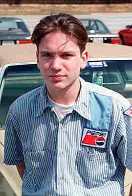 A picture of a white male youth with brown hair swept back and a blue-and-white-striped, short-sleeved button-up shirt with a pepsi logo above the left chest pocket. 