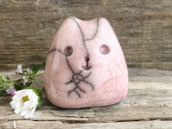 on a background of grey-brown driftwood, a cute little cat raku sculpture. it's glazed in pale pink and has a very happy face. its general shape is kind of round tapering up to two lil ears.
there are a daisy and a sprig of thyme (tiny green leave and tiny pink flowers) next to it on the left.