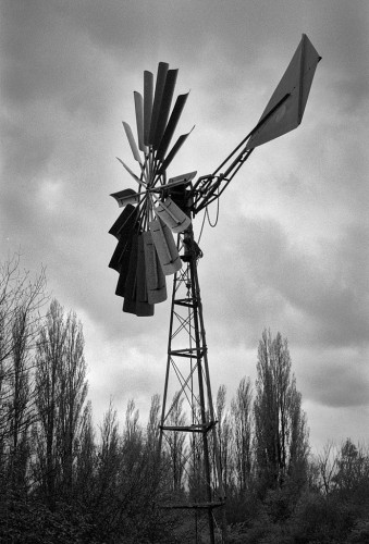 Black and white portrait format photo showing the dramatic silhouette of a wind pump against a moody sky. The wind pump has a three-sided conical metal tower, multiple large metal blades, here seen almost side on, and a large metal vane to keep it pointed into the wind. The lower part of the image shows distant bare winter Lombardy poplars.
